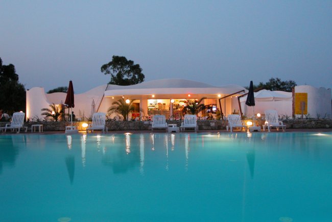 Piscina tranquilla al tramonto con sedie a sdraio e un edificio illuminato sullo sfondo.